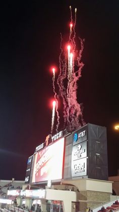 fireworks are lit up in the night sky above a stadium's scoreboard as people watch