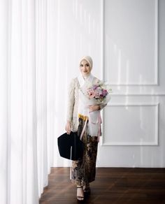 a woman standing in front of a white wall holding a bouquet of flowers and a black purse