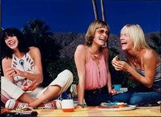 three women sitting on the ground laughing and eating food at an outdoor picnic with palm trees in the background