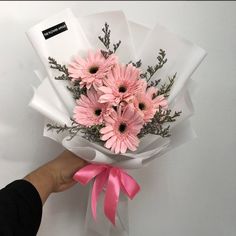 a person holding a bouquet of pink daisies in white paper with a pink ribbon