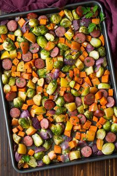 a pan filled with vegetables and sausages on top of a wooden table next to a purple towel