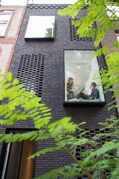 two people are sitting in the window of a brick building with green leaves around them