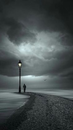 a person standing on the beach next to a light pole under a dark sky with clouds