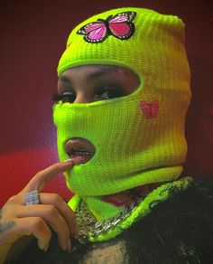 a woman wearing a neon green knitted mask and finger pointing at her nose with a butterfly on it