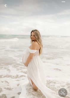 a pregnant woman standing in the surf at the beach with her arms around her waist