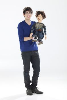 a man holding a doll in his right hand and smiling at the camera while standing against a white background