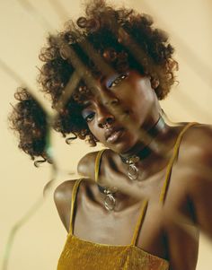 a woman with curly hair wearing a yellow top and necklaces looking at the camera