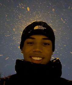 a man wearing a black hat and jacket in front of a blue sky with snow flakes