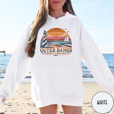 Celebrate the beauty of North Carolina's beautiful islands and coastline with our OBX Outer Banks Hoodie from Triangle Threads. This crewneck celebrates the beauty of the Cape Hatteras Lighthouse and the pristine sand dunes of the Outer Banks. Perfect for an OBX lover, or to remind you of that awesome summer vacation, this Gildan 18500 50/50 blend hooded sweatshirt comes in all sizes big and small for oversize and regular fits. So grab one today and celebrate the Carolina Coast and the beauty of Outer Banks Hoodies, White Hooded Hoodie For Beach Season, Hooded Hoodie For Vacation In Winter, Hooded Hoodie For Winter Vacation, Hooded Winter Hoodie For Vacation, White Long Sleeve Hoodie For Beach Season, White Long Sleeve Hoodie For Beach, Beach Season Long Sleeve Hoodie With Drawstring, Long Sleeve Hoodie For Beach In Fall