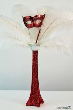 a red vase filled with white feathers and a masquerade mask on top of it