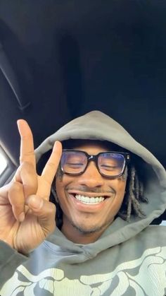 a man wearing glasses and a hoodie making the peace sign with his hand while sitting in a car