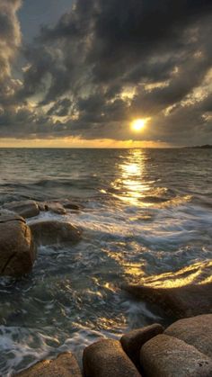 the sun is setting over the ocean with rocks in the foreground and dark clouds above