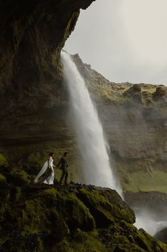 a man and woman holding hands standing in front of a waterfall