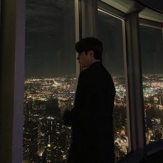 a man standing in front of a window looking out at the city lights and skyscrapers