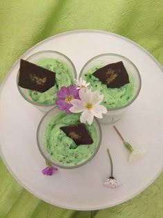 three small cups filled with green food on top of a white cake plate next to a flower