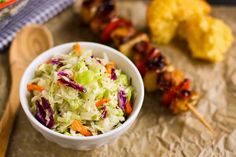 a white bowl filled with coleslaw next to skewers of meat and vegetables