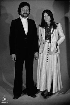 a man and woman standing next to each other in front of a white wall wearing long coats