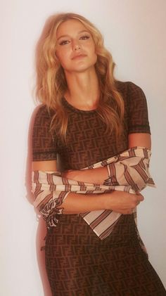 a woman with her arms crossed standing in front of a white wall wearing a brown and black dress