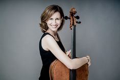 a woman is posing with her cello against a gray background and she has one arm on the back of the cello