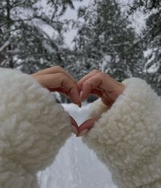 two hands making a heart shape with their fingers in front of snow covered trees and evergreens