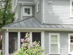 a gray house with white trim and some purple flowers in the foreground, on a sunny day