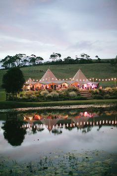 a group of tents set up next to a lake with lights on the grass and trees in the background