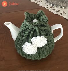 a knitted tea pot cover with white flowers on the front and green yarn in the back