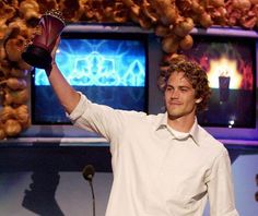 a young man holding up a trophy in front of two televisions and a microphone