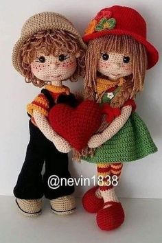 two knitted dolls holding a red heart and wearing matching hats, standing next to each other