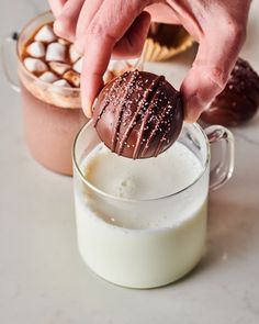 a person dipping a chocolate ball into a glass of milk with marshmallows in the background