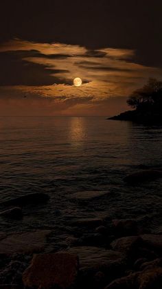 the moon is setting over the ocean with rocks