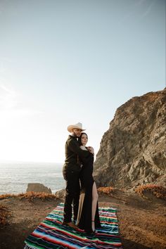 a man and woman standing on top of a blanket next to the ocean with their arms around each other