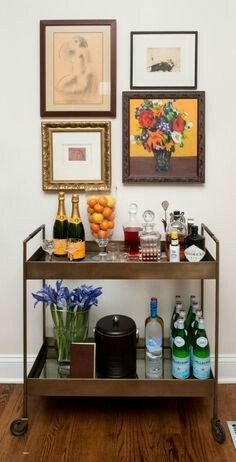 a table topped with bottles of wine next to pictures on the wall and vases filled with fruit