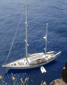 a sailboat floating in the ocean near some rocks and plants on the shore with blue water