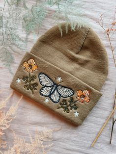 a brown hat with a butterfly embroidered on the front and side, sitting next to dried flowers