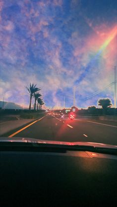 a rainbow is seen in the sky as cars drive down the road with palm trees on either side