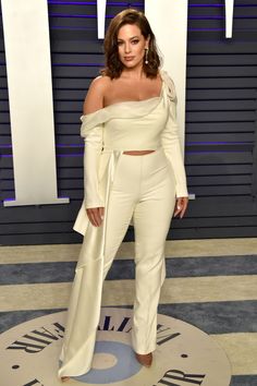 a woman standing in front of a wall wearing white pants and off the shoulder top