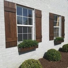 three windows with wooden shutters on the side of a white brick building and green bushes in front