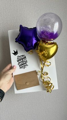 a hand is holding a card with balloons and streamers attached to it, next to an envelope