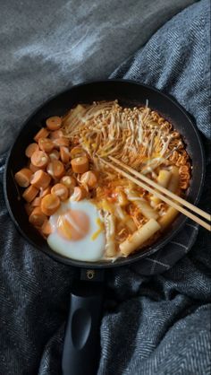 a skillet with noodles, eggs and chopsticks on a gray blanket next to a grey towel