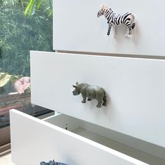 two toy zebras and a rhino on top of white drawers in front of a window