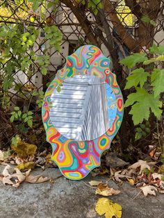 a colorful mirror sitting on the ground in front of a tree and fence with leaves around it