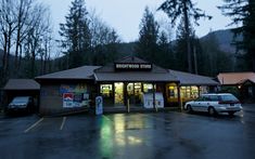 a small store with cars parked in the parking lot at night, next to trees and mountains