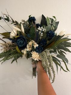 a hand holding a bouquet of flowers and greenery on a white wall behind it