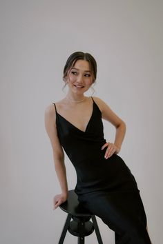 a woman in a black dress is sitting on a stool and posing for the camera