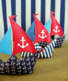 three paper boats with red sails are lined up against a striped wall, one is blue and the other has white anchors