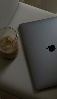 an apple laptop computer sitting on top of a white table next to a glass of liquid