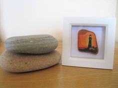 two rocks sitting on top of a wooden table next to a small white frame with a painting of a lighthouse