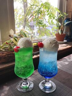 two glasses filled with different colored drinks on top of a table next to a window