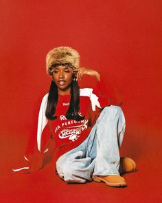 a woman sitting on the ground wearing a red shirt and fur hat with her legs crossed